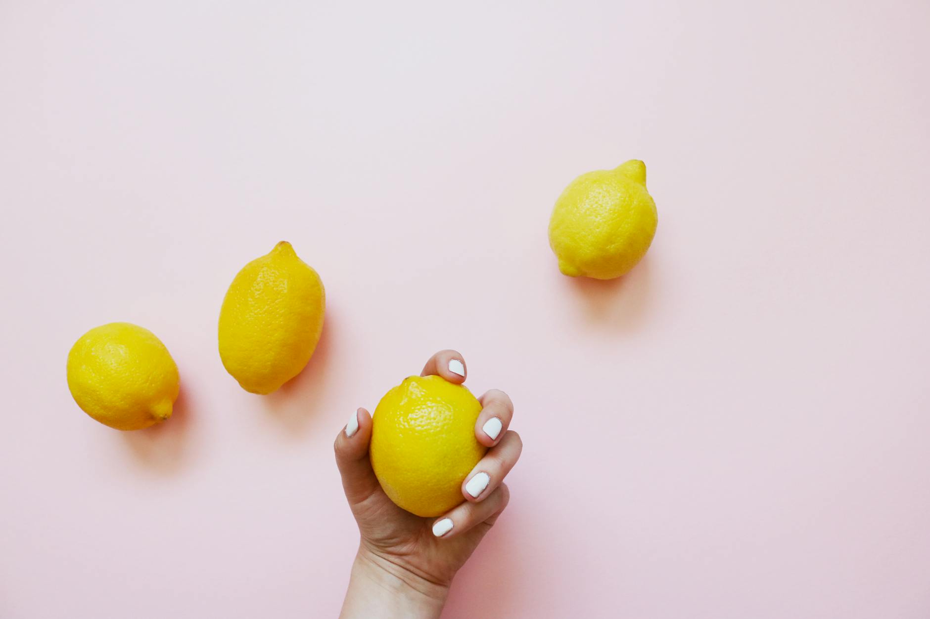 Eine Hand mit weißen Nägeln hält eine Zitrone vor einem weichen rosa Hintergrund, umgeben von drei weiteren Zitronen