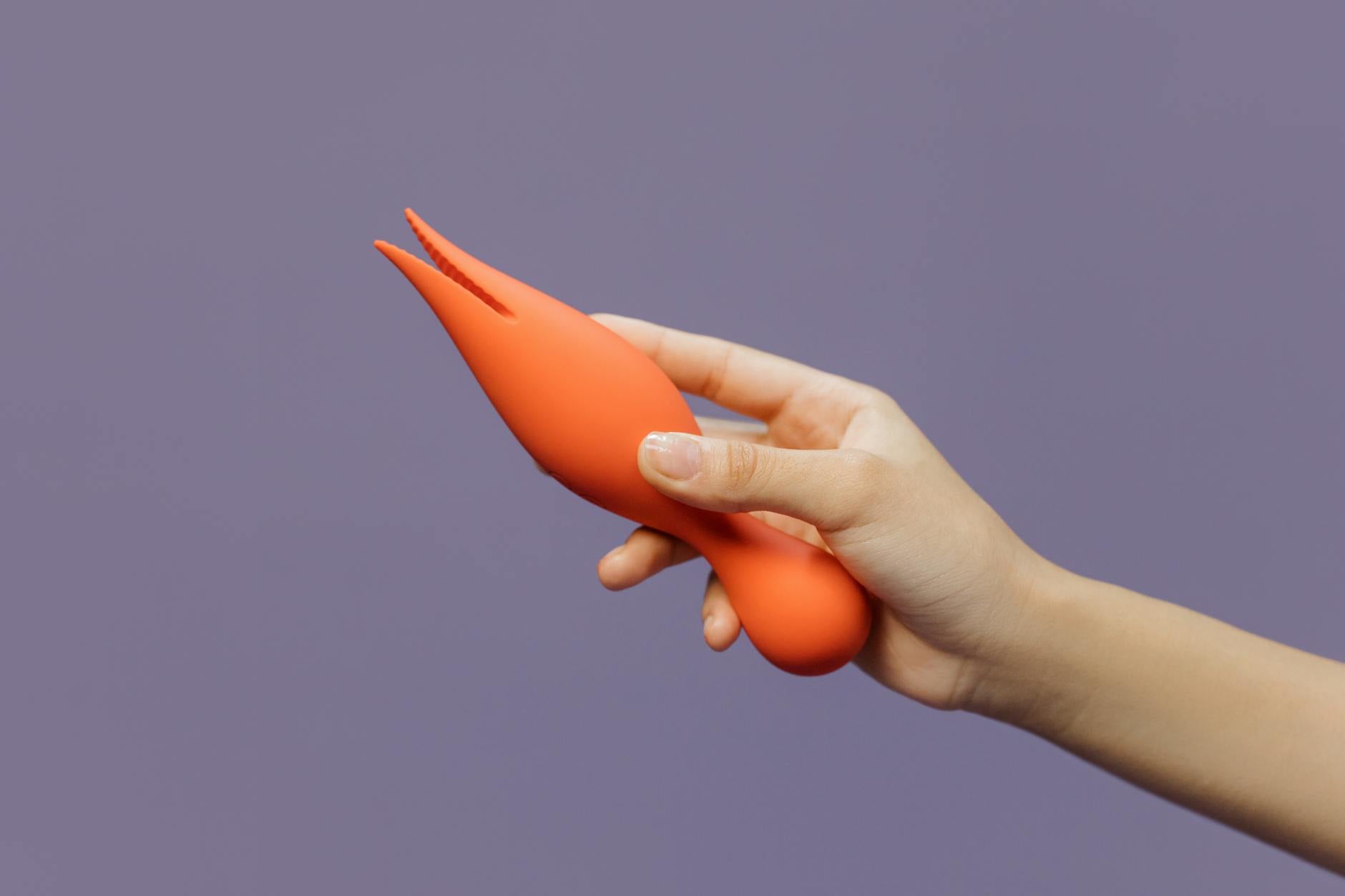 Nahaufnahme einer Hand, die einen orangen Vibrator gegen einen minimalistischen lila Hintergrund hält, der moderne Sinnlichkeit zeigt.