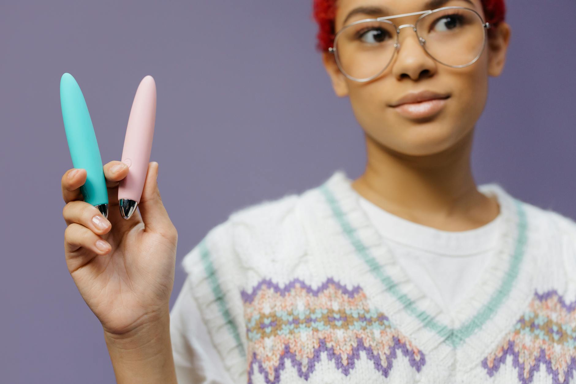 Junge Frau mit Brille hält zwei farbenfrohe Vibratoren gegen einen violetten Hintergrund