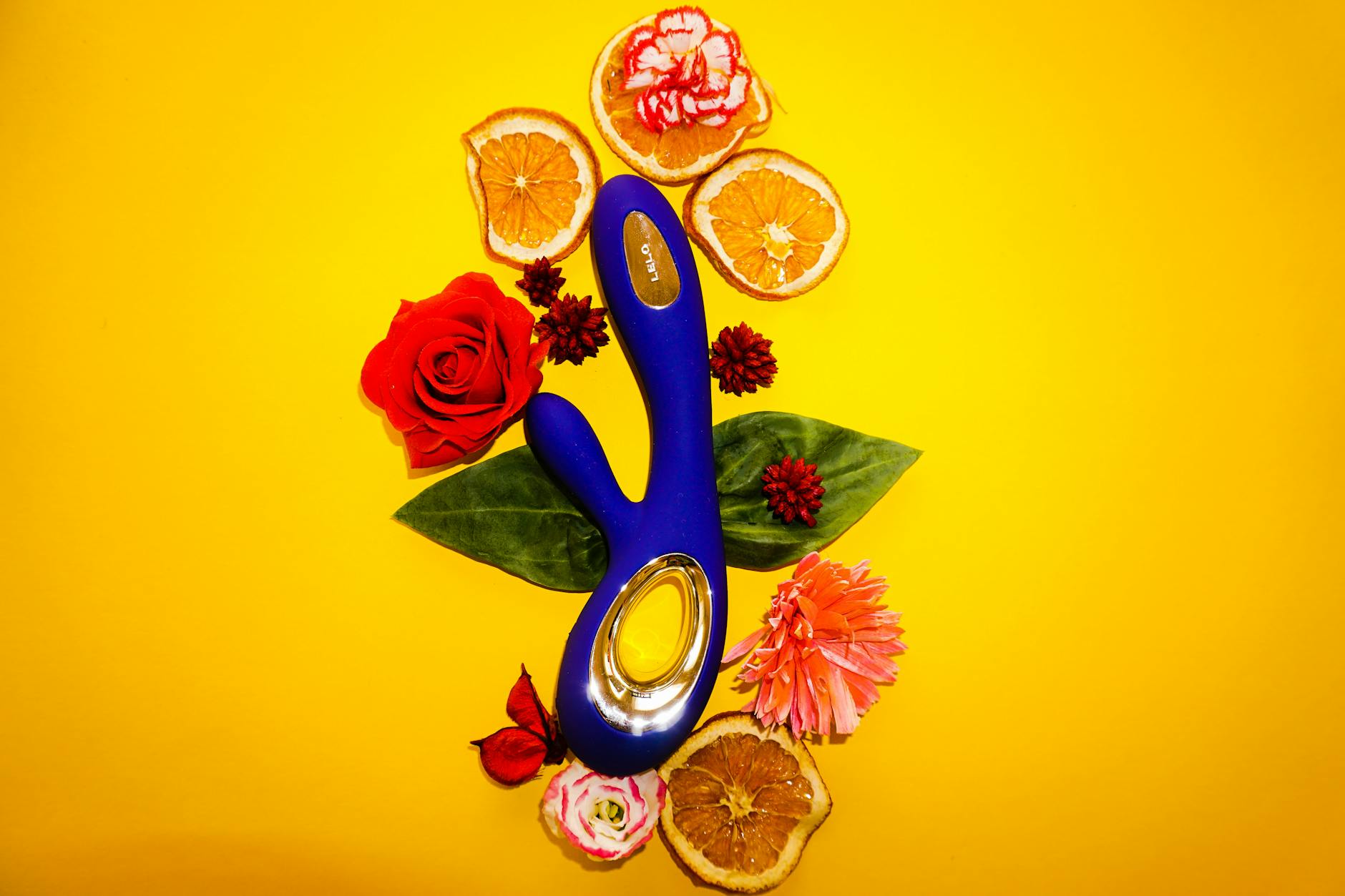 Blauer Lemon-Vibrator umgeben von Blumen und Orangenscheiben auf gelbem Hintergrund